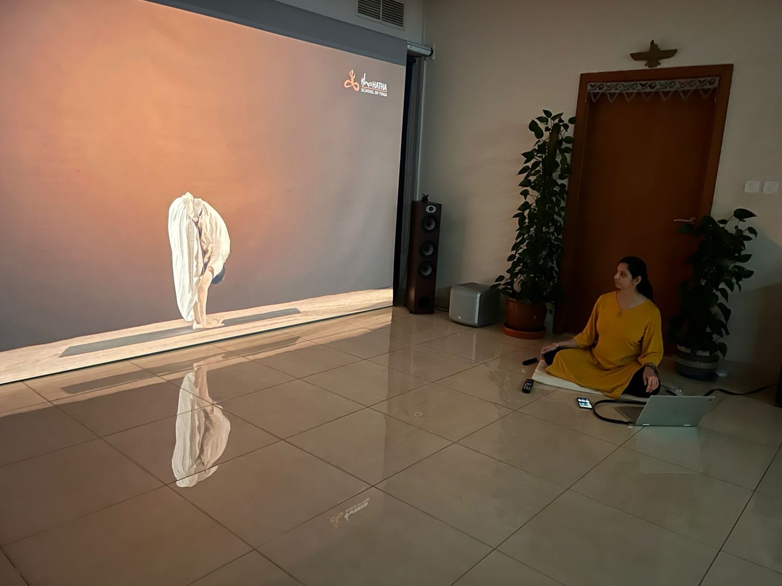The Yoga Quest studio: a bright tiled room with a large screen, plants and a bookshelf, Persis seated on a mat